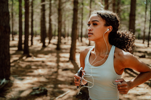 Смешанная раса спортсменка бегает в лесу, слушая музыку с наушниками - Фото, изображение