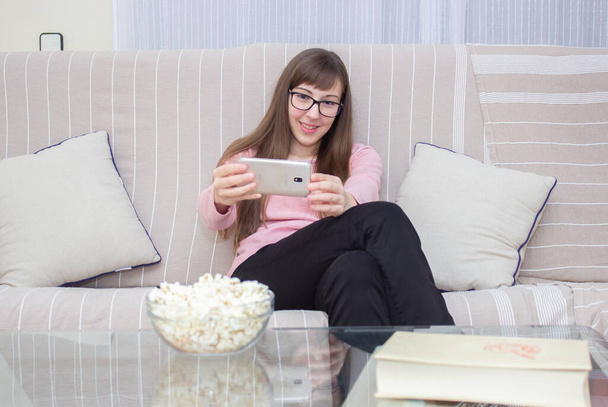 kobieta siedząca na kanapie wykonująca rozmowy wideo z telefonem i miską popcornu na stole - Zdjęcie, obraz