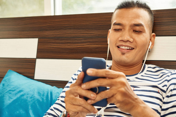 Podekscytowany przystojny młody Azjata słucha muzyki za pośrednictwem smartfona i SMS-a przyjaciela lub dziewczyny - Zdjęcie, obraz