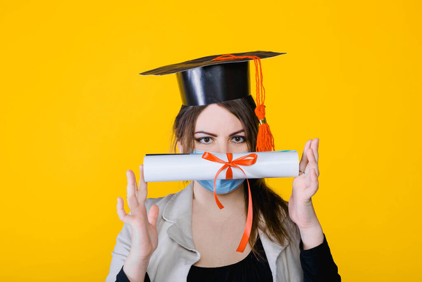 Graduate 2021. Portrait of young brunette woman in face medical mask and graduate hat in studio with yellow isolated background - Φωτογραφία, εικόνα