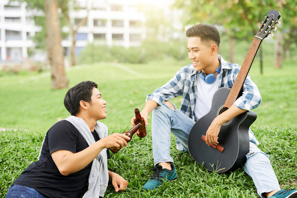 Two young men sitting on green grass with guitar and drinking beer outdoors - Photo, Image