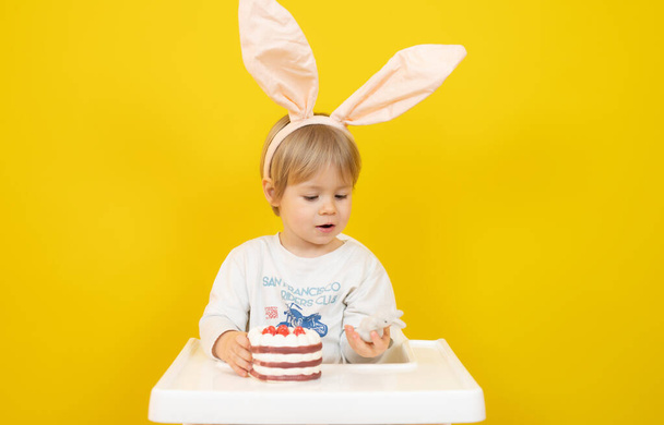 Hermoso bebé con diadema de conejo sentado y sosteniendo un pastel aislado sobre fondo amarillo. - Foto, imagen