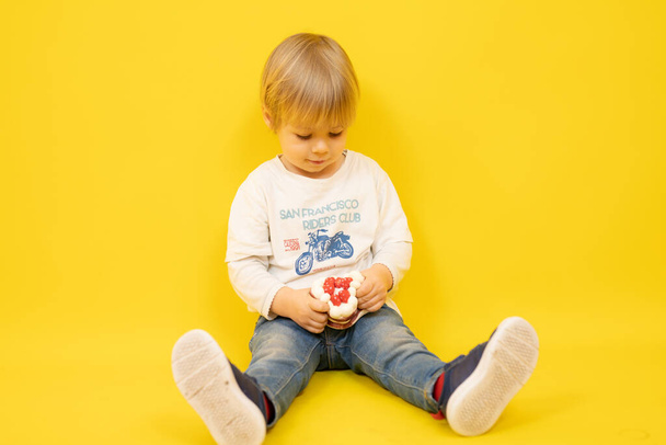 Beautiful baby boy sitting and holding a cake isolated over yellow background. - Zdjęcie, obraz