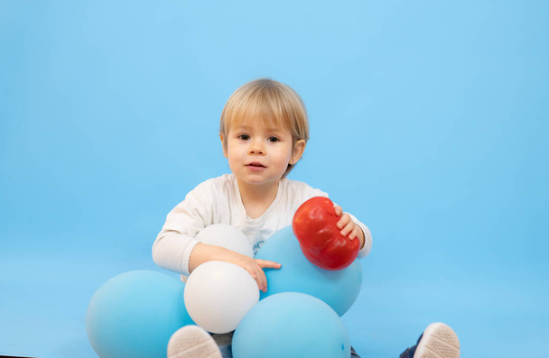 Piękny uśmiechnięty chłopiec siedzi z balonami odizolowane na niebieskim tle. - Zdjęcie, obraz