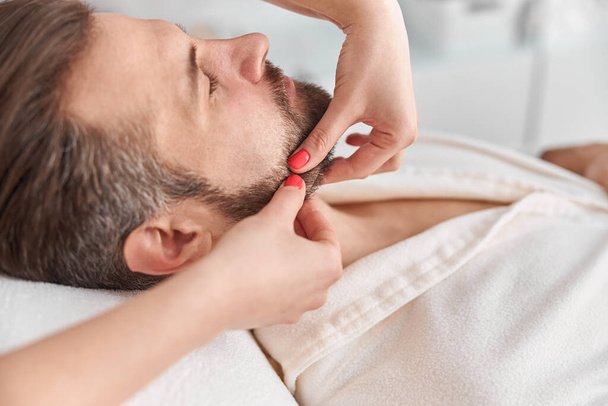 O homem com barba está deitado de costas, a fazer massagens no rosto. Tratamento de beleza de massagem facial. Conceito de bem-estar, beleza e relaxamento - Foto, Imagem