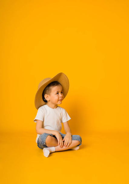 malý chlapec sedí v tričku a šortkách se slamákem a dívá se stranou na žluté pozadí - Fotografie, Obrázek