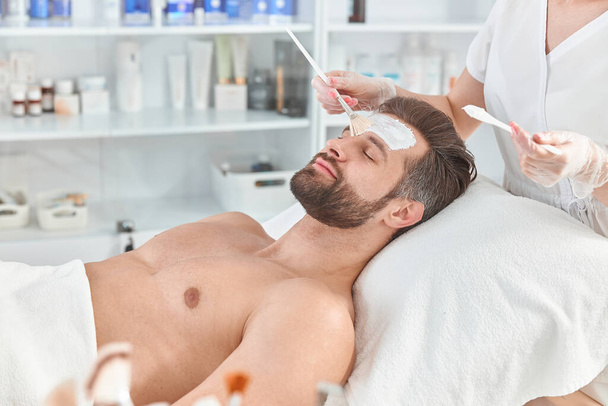 Бородатий молодий чоловік розслабляється, поки косметолог розкладає білу глину на обличчі
 - Фото, зображення