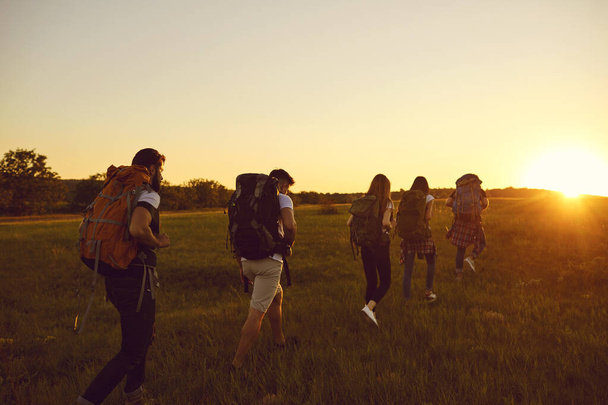 Group of five people with large backpacks move in a row across a field at sunset. - Photo, image