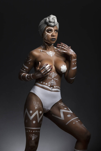 Афроамериканка только в нижнем белье, с краской для тела, макияжем, шарфом на голове, и руками ее обнаженное тело, студия с серым фоном - Фото, изображение
