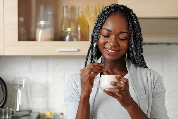 Portret uśmiechniętej młodej czarnej kobiety mieszającej mleko z cukrem w filiżance porannej kawy - Zdjęcie, obraz