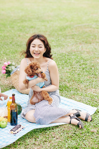 Parkta battaniyede oturup sevimli köpeğiyle oynayan mutlu, heyecanlı genç bir kadın. - Fotoğraf, Görsel