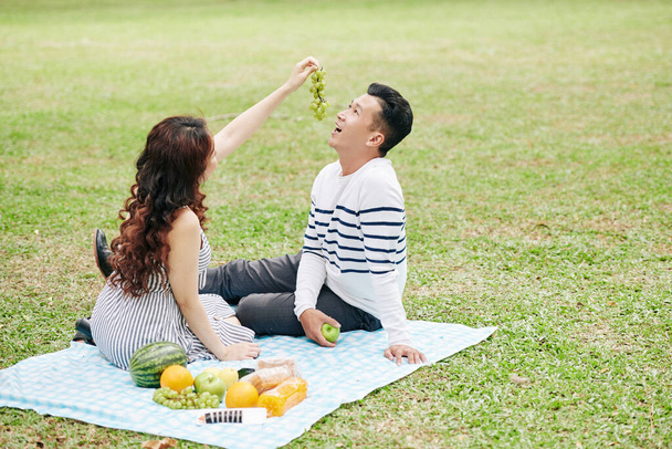 Jolie jeune femme nourrissant petit ami avec des raisins quand ils sont assis sur la couverture dans le parc de la ville - Photo, image