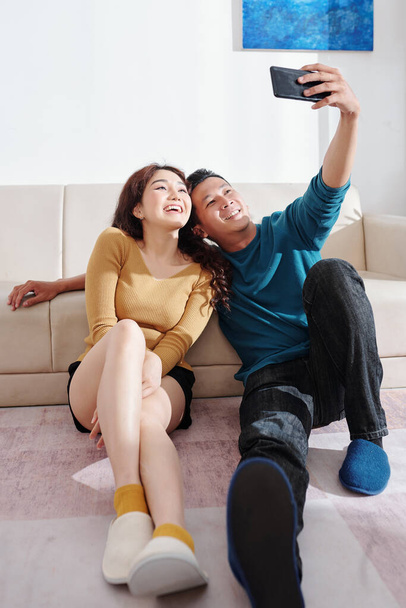 Fröhliches junges vietnamesisches Paar verbringt Wochenende zu Hause, sitzt auf dem Boden und macht gemeinsam Selfie - Foto, Bild