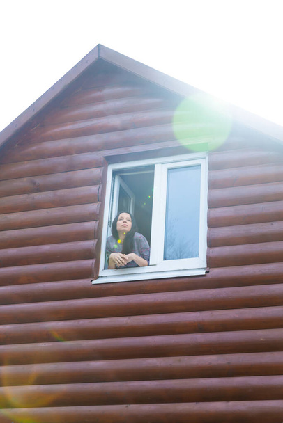 Το κορίτσι κοιτάζει έξω από το παράθυρο του δεύτερου ορόφου ενός εξοχικού. - Φωτογραφία, εικόνα