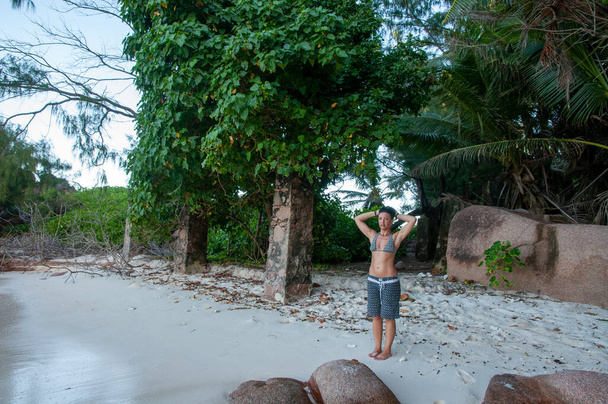 Anse Cayman, La Digue island, Σεϋχέλλες Μια νεαρή τουρίστρια στέκεται στην παραλία με πυκνή βλάστηση στο βάθος - Φωτογραφία, εικόνα