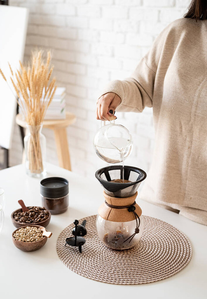 Alternative coffee brewing. Young woman brewing coffee in coffee pot standing at the white table with various stuff for alternative coffee brewing, pouring hot water into the filter - 写真・画像