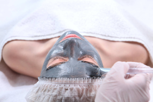 A cosmetologist applies a cosmetic mask to a woman's face.The woman's face is covered with an alginate mask. Cosmetic procedure. A gray, frozen mass on the woman's face.Light background.Copy space .Concept of beauty. - Foto, imagen