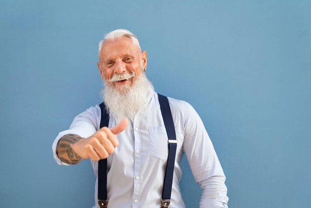 Fashionable senior man with grey beard doing happy thumbs up gesture with hand, looking at the camera and smiling. Blue pastel background. - Foto, imagen