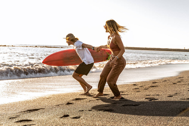 Beautiful happy surfer couple having fun on the sandy beach, running with surfboard to the waves. Summer vibes. Sunset light.  - Fotó, kép