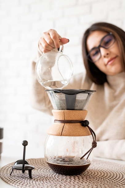 Alternativní vaření kávy. Mladá žena vaří kávu v kávové konvice stojící u bílého stolu s různými věcmi pro alternativní vaření kávy, nalévání horké vody do filtru - Fotografie, Obrázek
