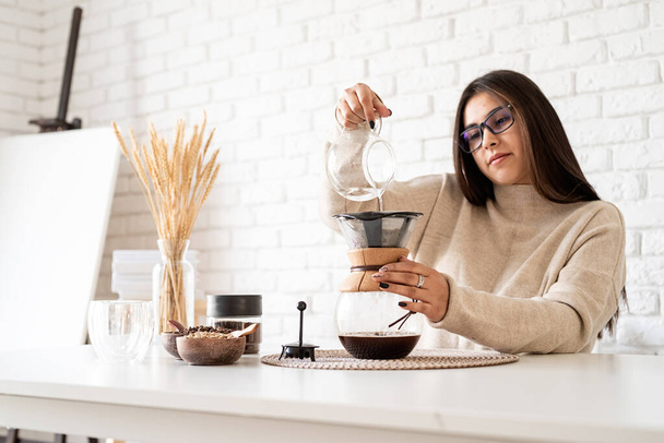 Alternatywne parzenie kawy. Młoda kobieta parzy kawę w dzbanku do kawy stojąc przy białym stole z różnymi rzeczami do alternatywnego zaparzania kawy, wlewając gorącą wodę do filtra - Zdjęcie, obraz