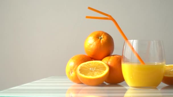 Jugo de naranja en un vaso rodeado de naranjas naturales con un fondo blanco claro al sol natural - Imágenes, Vídeo