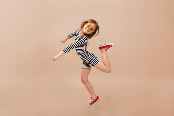 Full-leght φωτογραφία του μικρού όμορφου κοριτσιού με ριγέ φόρεμα και κόκκινα sneakers που χορεύουν και πηδούν πάνω από μπεζ φόντο. Υψηλής ποιότητας φωτογραφία - Φωτογραφία, εικόνα