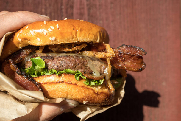 Una mano sosteniendo una fresca y jugosa hamburguesa de barbacoa occidental con queso, frito, cebolla, lechuga, tocino, salsa barbacoa en un bollo de semillas de sésamo frente a un fondo rojo. - Foto, imagen