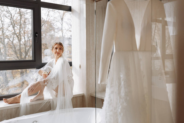 Brautmorgen. Braut trinkt Champagner im Peignoir. Die junge Frau sitzt auf einem großen Fenster in einem Hotelzimmer im Badezimmer. Schöne Mädchen in weißer Hochzeitsrobe. Hochzeitstag. - Foto, Bild