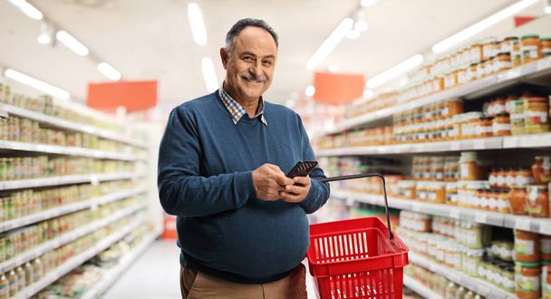 Mature man holding a shopping basket and a mobile phone inside a supermarket and looking at camera - Zdjęcie, obraz