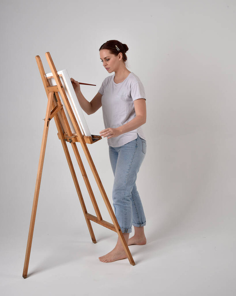 Retrato de comprimento total de uma menina artista de cabelos vermelhos vestindo jeans casuais e camisa branca. pose de pé pintando uma tela em um cavalete, contra um fundo de estúdio. - Foto, Imagem