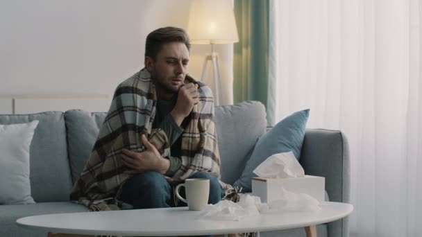 Mladý nemocný chlapík zabalený v kostkované trpí záchvat kašle a pije uzdravující čaj, sedí sám na gauči doma - Záběry, video