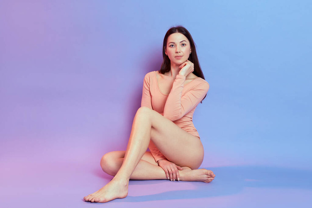 Attractive girl with perfect body sitting on floor and holding chin with fist, looks at camera with serious expression, dresses beige bodysuit, isolated over blue background with pink neon light. - Фото, изображение