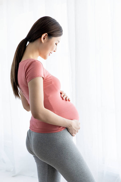 Side view of beautiful asian pregnant woman standing near window at home - Photo, Image