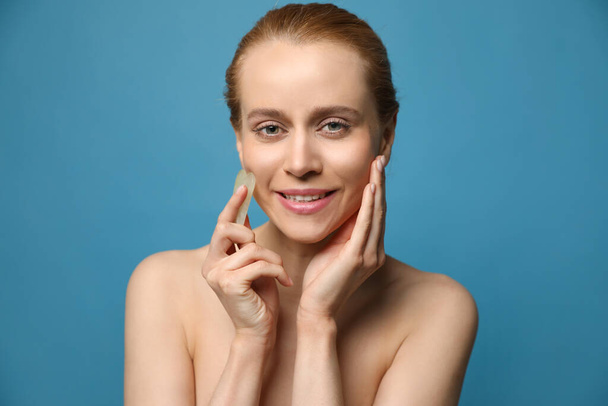 Beautiful young woman doing facial massage with gua sha tool on blue background - Fotoğraf, Görsel