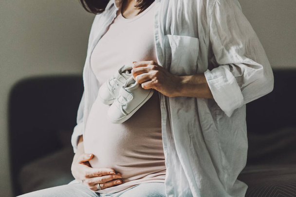 Pregnant woman holding small baby shoes. Lifestyle. Mothernity concept.  - Fotó, kép