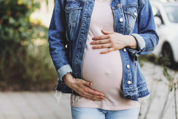 Pregnant woman in jeans jacket standing outdoors and touching belly.  - Fotografie, Obrázek