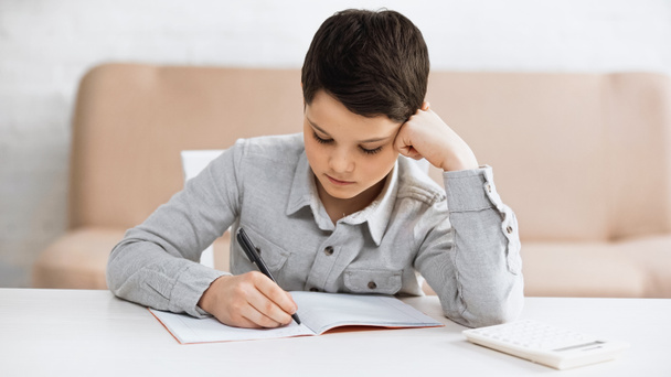 Junge schreibt auf Notizbuch neben Taschenrechner auf Tisch zu Hause  - Foto, Bild