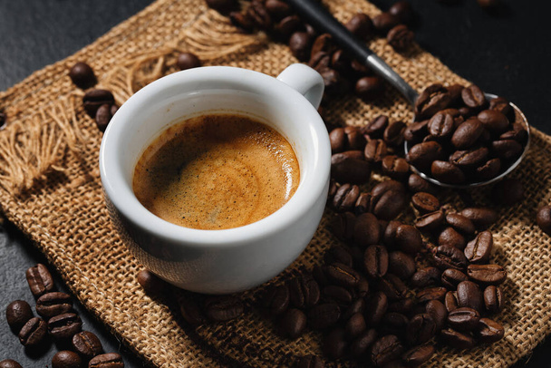 Tasty espresso served in cup with coffee beans around and spoon. Closeup. Dark background.  - Photo, Image