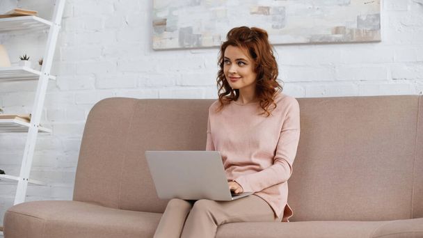Smiling woman using laptop on couch in living room  - Foto, Bild
