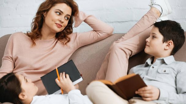 Woman looking at kids with notebook and book on blurred foreground on couch  - Фото, изображение