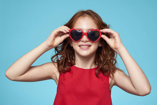 Dziewczynka w okularach przeciwsłonecznych w postaci serc czerwona sukienka kręcone włosy niebieskie tło - Zdjęcie, obraz