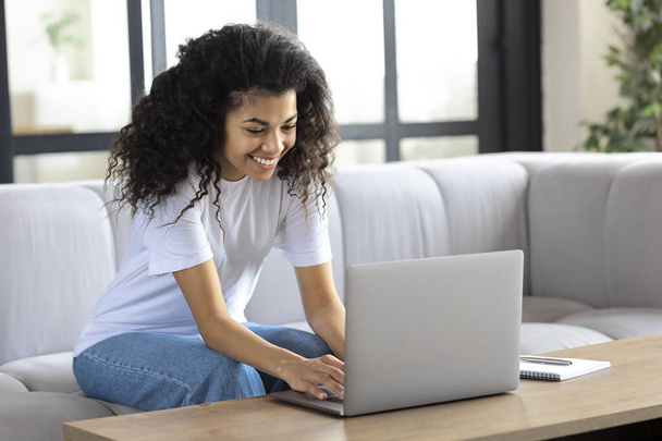 Young successful african american woman sitting at home on the couch streaming something on a laptop - Photo, Image