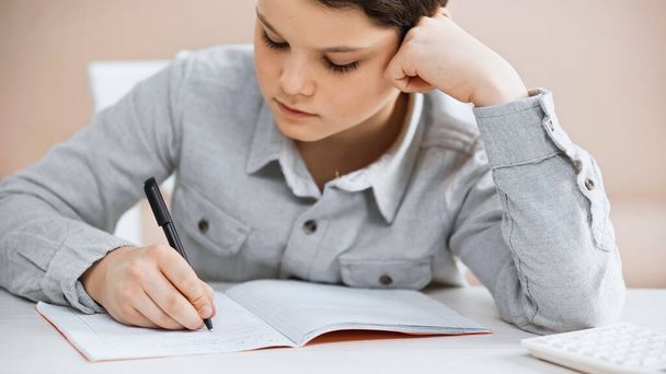 Preteen boy writing on notebook near burred calculator at home  - Foto, Bild