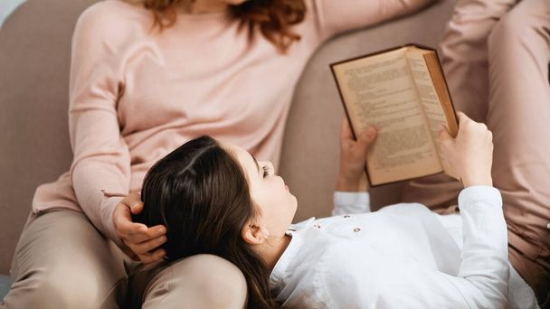 Girl reading book near mother on blurred background on couch  - Photo, image