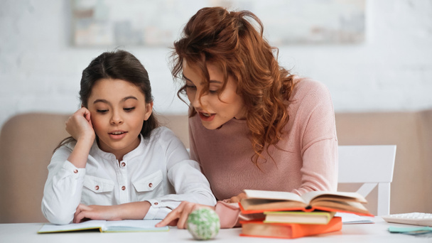 Frau spricht mit Tochter, während sie in der Nähe von Büchern bei Hausaufgaben hilft  - Foto, Bild