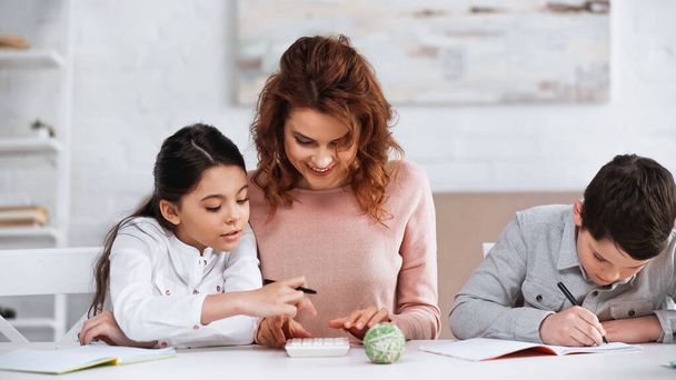 Smiling woman sitting near daughter using calculator and son writing on notebook  - Foto, Bild
