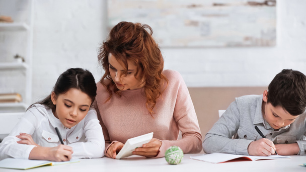 Woman holding calculator near kids writing on notebooks at home  - Foto, Bild
