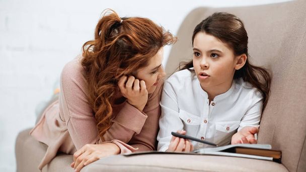 Menina falando perto de mãe e laptop ao fazer lição de casa no sofá  - Foto, Imagem