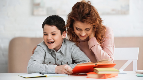 Smiling woman sitting near cheerful son holding book on blurred foreground  - Fotografie, Obrázek